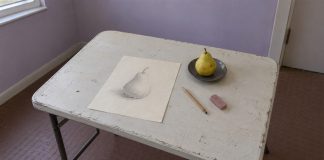 A graphite pear sketch, pencil and pink eraser beside a pear on a gray saucer, on a worn folding table in a lilac room.