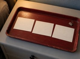 Three blank lined index cards and a steel binder clip on a worn dark-red tray beside a bed in warm light.