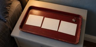 Three blank lined index cards and a steel binder clip on a worn dark-red tray beside a bed in warm light.
