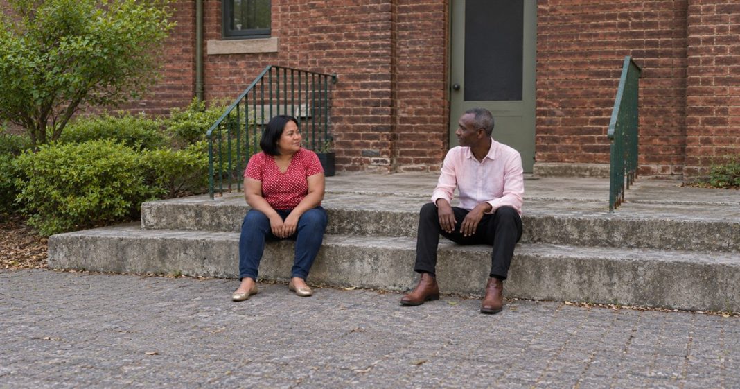 Relationship clarity — courtyard step conversation A woman in a red polka-dot blouse and a man in a pale pink shirt talking while seated apart on concrete courtyard steps beside green railings.
