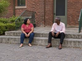 A woman in a red polka-dot blouse and a man in a pale pink shirt talking while seated apart on concrete courtyard steps beside green railings.