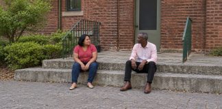 A woman in a red polka-dot blouse and a man in a pale pink shirt talking while seated apart on concrete courtyard steps beside green railings.