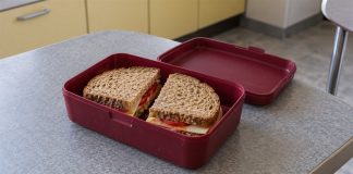 A brown-bread cheese and tomato sandwich in an open burgundy lunchbox with its lid beside it on a gray kitchen table.