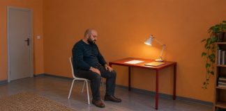 A bald, bearded man in a striped shirt seated with hands on his thighs beside a red desk, blank paper and a lit task lamp in an orange-walled room.