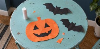 An orange smiling paper pumpkin, two black paper bats, a capped glue stick and paper scraps on a worn turquoise craft table.