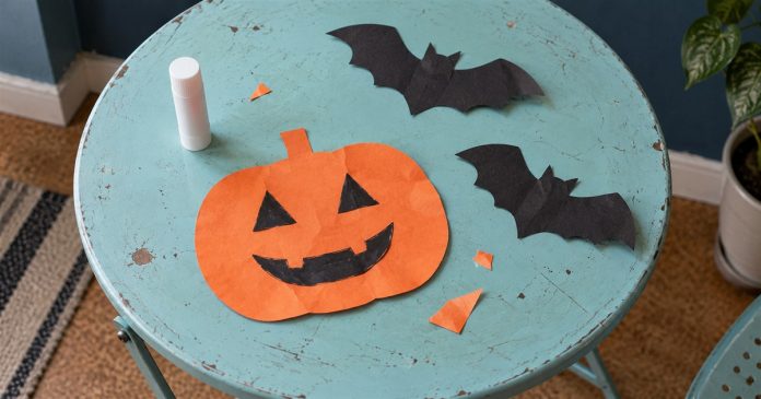 An orange smiling paper pumpkin, two black paper bats, a capped glue stick and paper scraps on a worn turquoise craft table.