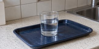 A clear drinking glass about half filled with water on a scratched navy tray, beside a kitchen sink on a cream speckled counter.