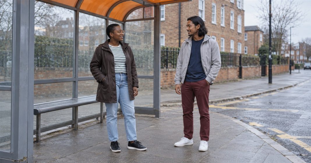 2292 realistic bus shelter conversation 20261005 A short-haired woman in amber glasses and a brown raincoat talking with a long-haired man in a gray jacket beside an orange-roofed bus shelter.