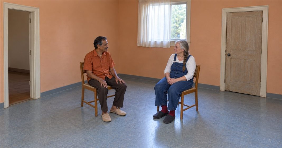 2299 realistic ordinary seated discussion 20261005 A mustached man in an orange shirt and a gray-haired woman in denim overalls talking from separate wooden cane chairs in an apricot-walled room.