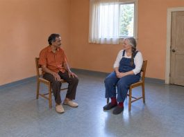A mustached man in an orange shirt and a gray-haired woman in denim overalls talking from separate wooden cane chairs in an apricot-walled room.