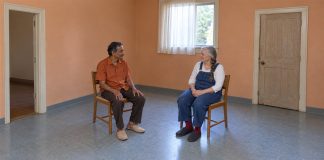A mustached man in an orange shirt and a gray-haired woman in denim overalls talking from separate wooden cane chairs in an apricot-walled room.