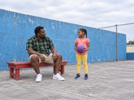 A man in a green checked shirt sitting on a red bench beside a girl in a coral shirt and yellow leggings holding a purple ball on a blue-walled court.