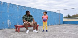 A man in a green checked shirt sitting on a red bench beside a girl in a coral shirt and yellow leggings holding a purple ball on a blue-walled court.