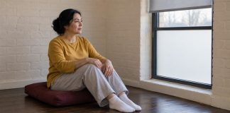 A woman in a mustard-yellow shirt and light gray trousers sitting on a maroon floor cushion beside a frosted window in a cream brick room.
