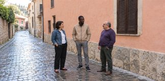 A woman in a denim jacket and two men in tan and purple tops talking on a cobblestone lane beside a salmon-colored wall and brown shutters.