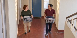 A red-haired woman in a green sweater and a mustached man in a burgundy polo carrying separate gray laundry baskets in a cream hallway with red tiles and a blue door.