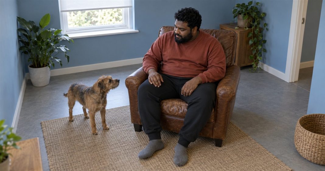Breakup — Quiet Pet Company — Realistic Cover 20261005 A man in a rust-red sweatshirt sitting in a brown leather armchair and looking toward a small brown terrier on a woven rug in a blue-walled room.