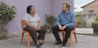An older woman in a gray striped shirt and a blond man in a blue hoodie talking from separate orange chairs in a paved courtyard beside a lavender wall.