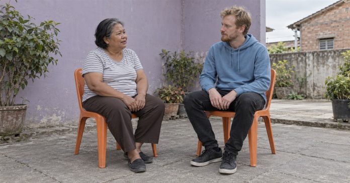 An older woman in a gray striped shirt and a blond man in a blue hoodie talking from separate orange chairs in a paved courtyard beside a lavender wall.