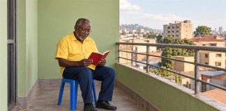 An older man in a yellow shirt and dark blue trousers reading a red book on a blue stool beside a metal balcony railing and a pale green wall.