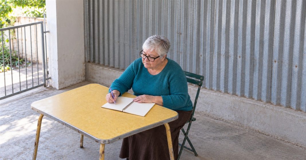 Relationship reflection — personal notes — realistic cover 2383 An older woman with silver hair and black glasses writing in a spiral notebook with a lavender pen at a yellow table beside a corrugated-metal wall.