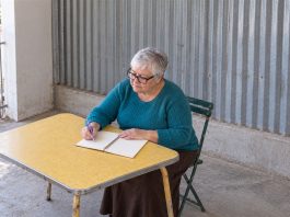 An older woman with silver hair and black glasses writing in a spiral notebook with a lavender pen at a yellow table beside a corrugated-metal wall.