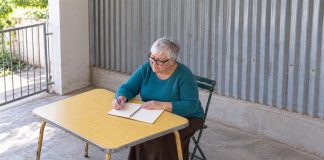 An older woman with silver hair and black glasses writing in a spiral notebook with a lavender pen at a yellow table beside a corrugated-metal wall.