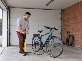 A man in a gray shirt and maroon trousers using a yellow floor pump beside a teal bicycle in a brick-walled garage with an open entrance.