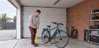 A man in a gray shirt and maroon trousers using a yellow floor pump beside a teal bicycle in a brick-walled garage with an open entrance.