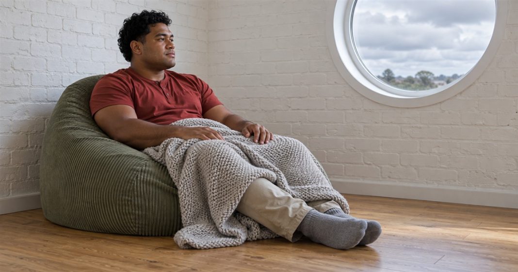 Realistic optimism — an ordinary restful pause A man in a red shirt resting on a green corduroy beanbag with a gray knitted blanket over his knees, beside a round window in a white brick room.