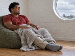 A man in a red shirt resting on a green corduroy beanbag with a gray knitted blanket over his knees, beside a round window in a white brick room.