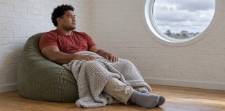 A man in a red shirt resting on a green corduroy beanbag with a gray knitted blanket over his knees, beside a round window in a white brick room.