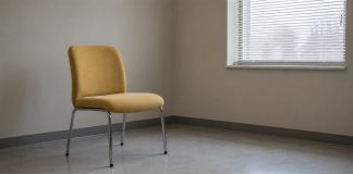 An empty yellow upholstered chair with chrome legs in a plain beige-gray room beside a window with white blinds.
