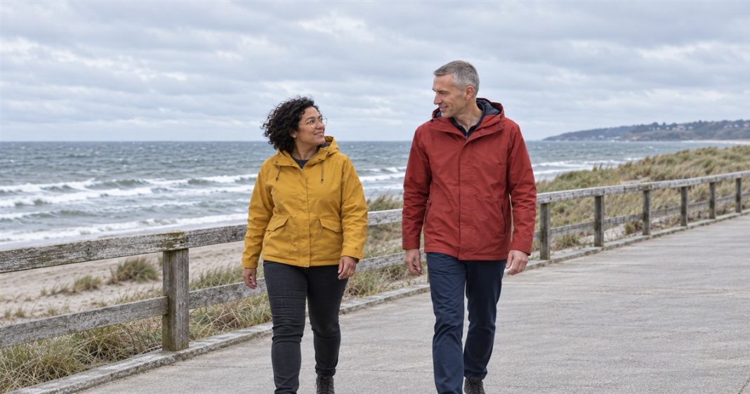 Twin flames meaning — ordinary seaside companionship A woman in a mustard-yellow jacket and a man in a brick-red raincoat talking while walking beside a wooden railing on an overcast seaside promenade.