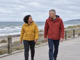 A woman in a mustard-yellow jacket and a man in a brick-red raincoat talking while walking beside a wooden railing on an overcast seaside promenade.