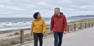 A woman in a mustard-yellow jacket and a man in a brick-red raincoat talking while walking beside a wooden railing on an overcast seaside promenade.