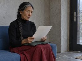 A woman with a gray-streaked braid reading a sheet of paper above a green folder on her lap, seated on a blue sofa beside a rain-streaked glass door.