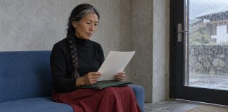 A woman with a gray-streaked braid reading a sheet of paper above a green folder on her lap, seated on a blue sofa beside a rain-streaked glass door.