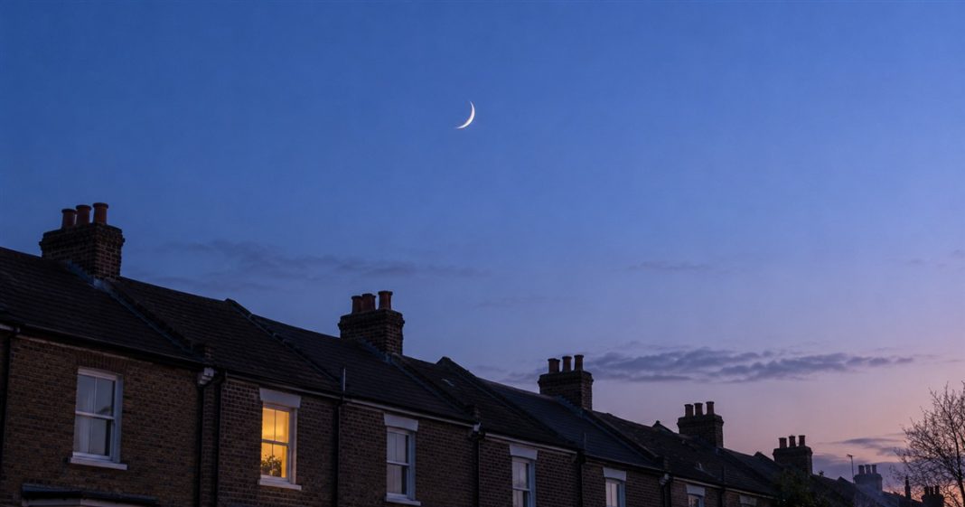 A slender crescent moon in a blue twilight sky above brick terraced-house rooftops and chimney stacks, with one warmly lit window.