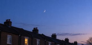 A slender crescent moon in a blue twilight sky above brick terraced-house rooftops and chimney stacks, with one warmly lit window.