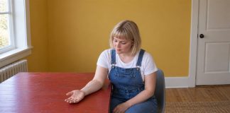 A woman in denim overalls and a white shirt looking at her relaxed open palm on a red wooden desk in a yellow-walled room.