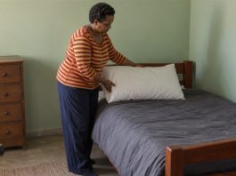 A woman in a red-and-yellow striped shirt placing a white pillow on a wooden bed with gray bedding in a pale-green room.
