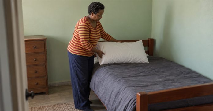 2459 Realistic small tidying step 20261005 A woman in a red-and-yellow striped shirt placing a white pillow on a wooden bed with gray bedding in a pale-green room.