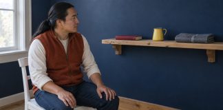 A man in a rust-orange vest seated on a white chair looking at a red book, yellow jug and folded gray cloth on a wooden shelf.