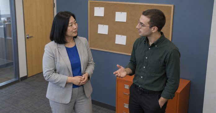 A woman in a gray suit and a man in a green shirt talking beside a corkboard and orange filing cabinet in an office.