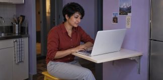 A woman in a brick-red shirt using a white laptop at a cream folding desk beside a violet kitchenette wall.