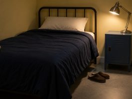 A single bed with navy bedding beside a lit metal lamp on a blue bedside locker, with brown slippers on the floor.