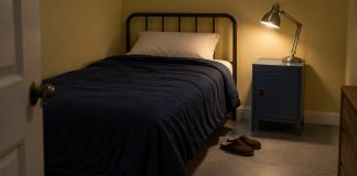 A single bed with navy bedding beside a lit metal lamp on a blue bedside locker, with brown slippers on the floor.