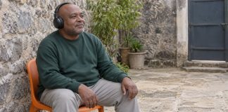 A man wearing black headphones and a green sweatshirt seated on an orange chair in a stone courtyard.