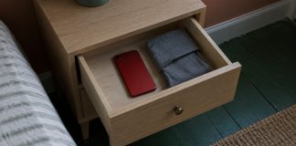 A red phone lying face down beside a folded gray pouch inside an open pale-oak bedside drawer.